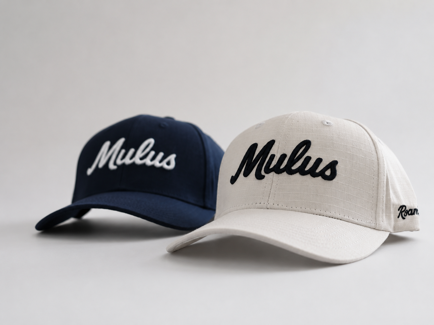 Two baseball caps on a light gray studio background: one navy blue with white embroidered 'Mulus' script and one off-white with black embroidered 'Mulus' script. A small side logo is visible on the brim of the front cap.