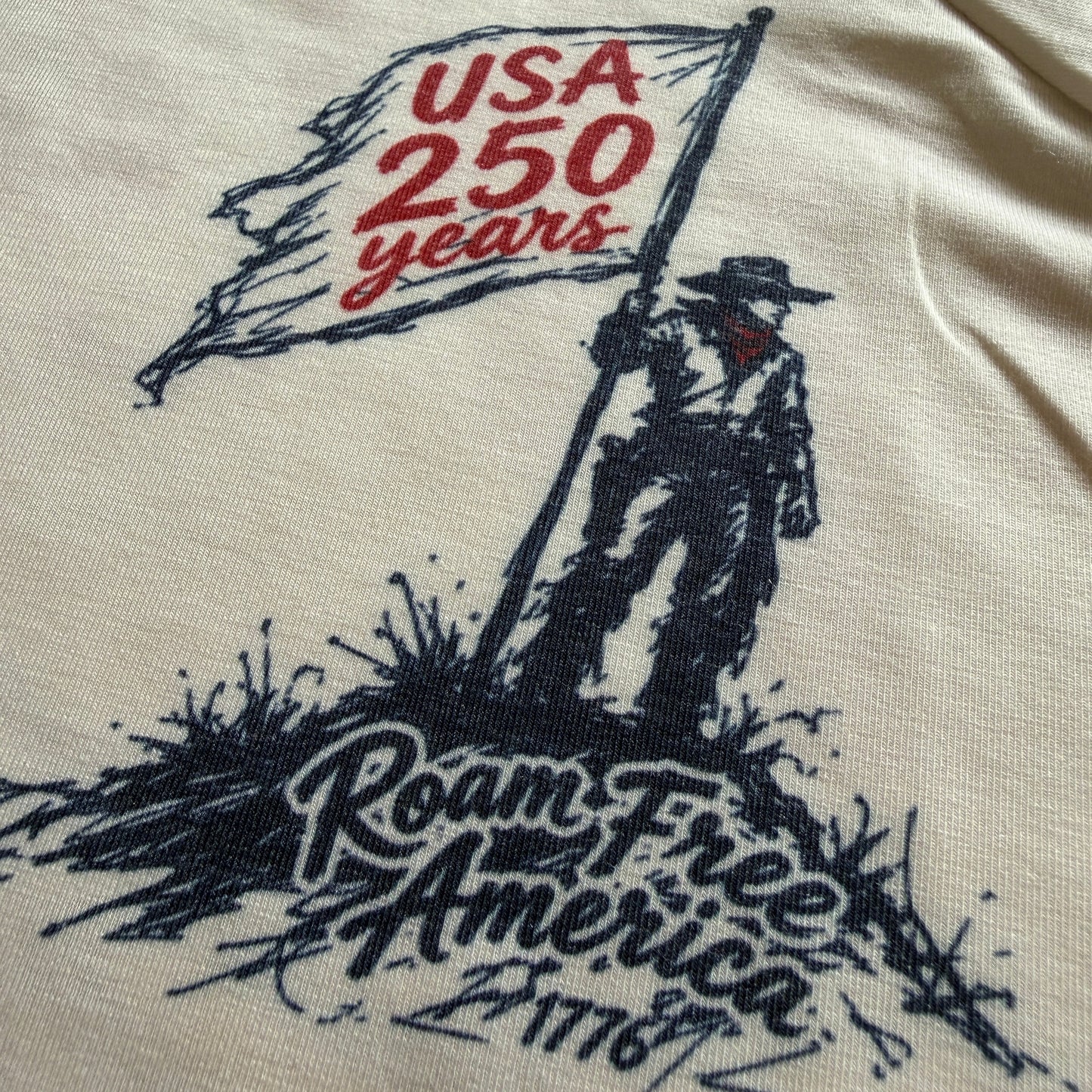 Close-up of a cream-colored t-shirt featuring a graphic print of a pioneer holding a flag that reads 'USA 250 years' and 'Roam Free America 1776'.