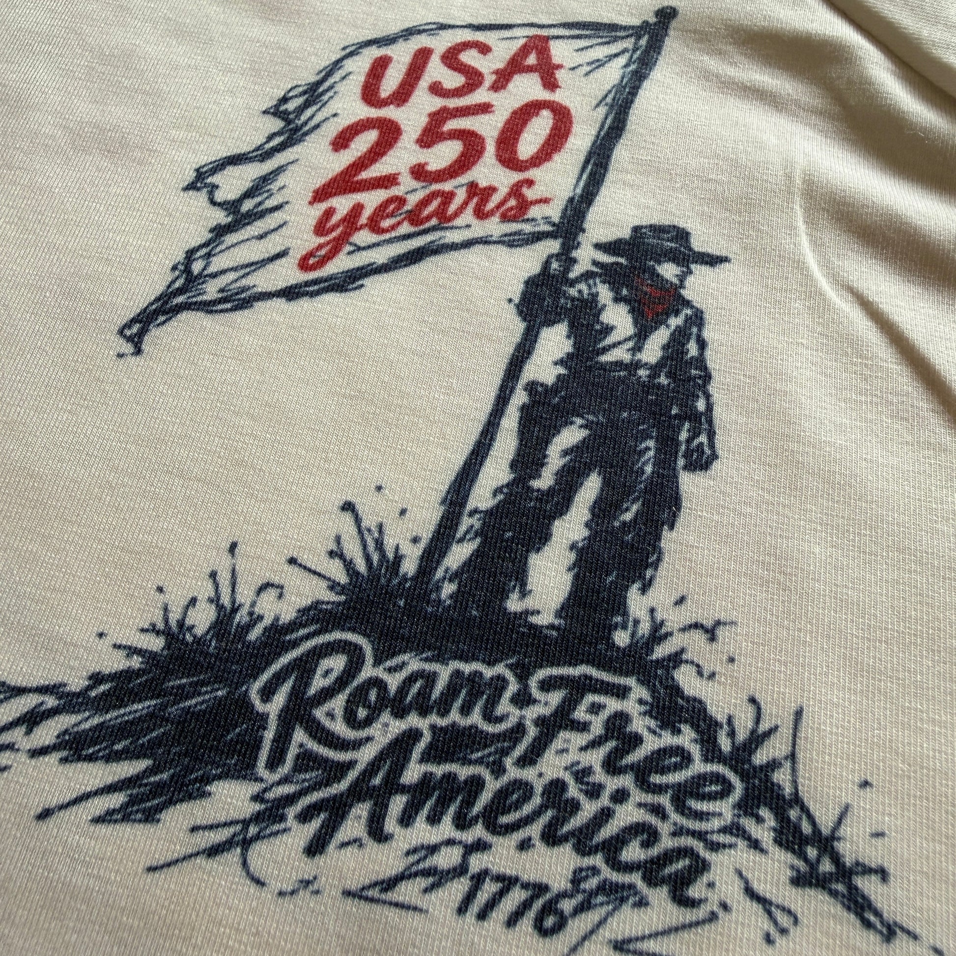 Close-up of a cream-colored t-shirt featuring a graphic print of a pioneer holding a flag that reads 'USA 250 years' and 'Roam Free America 1776'.