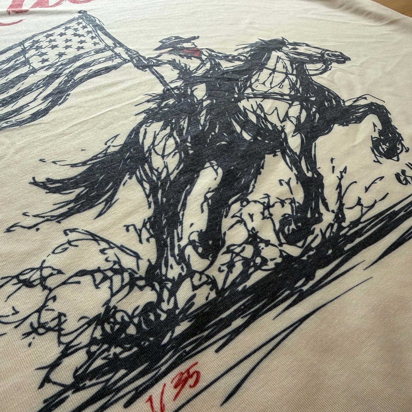 A close-up of a cream-colored t-shirt featuring a black ink sketch of a horse and rider, with a partial red script logo on the left and a red signature on the right.