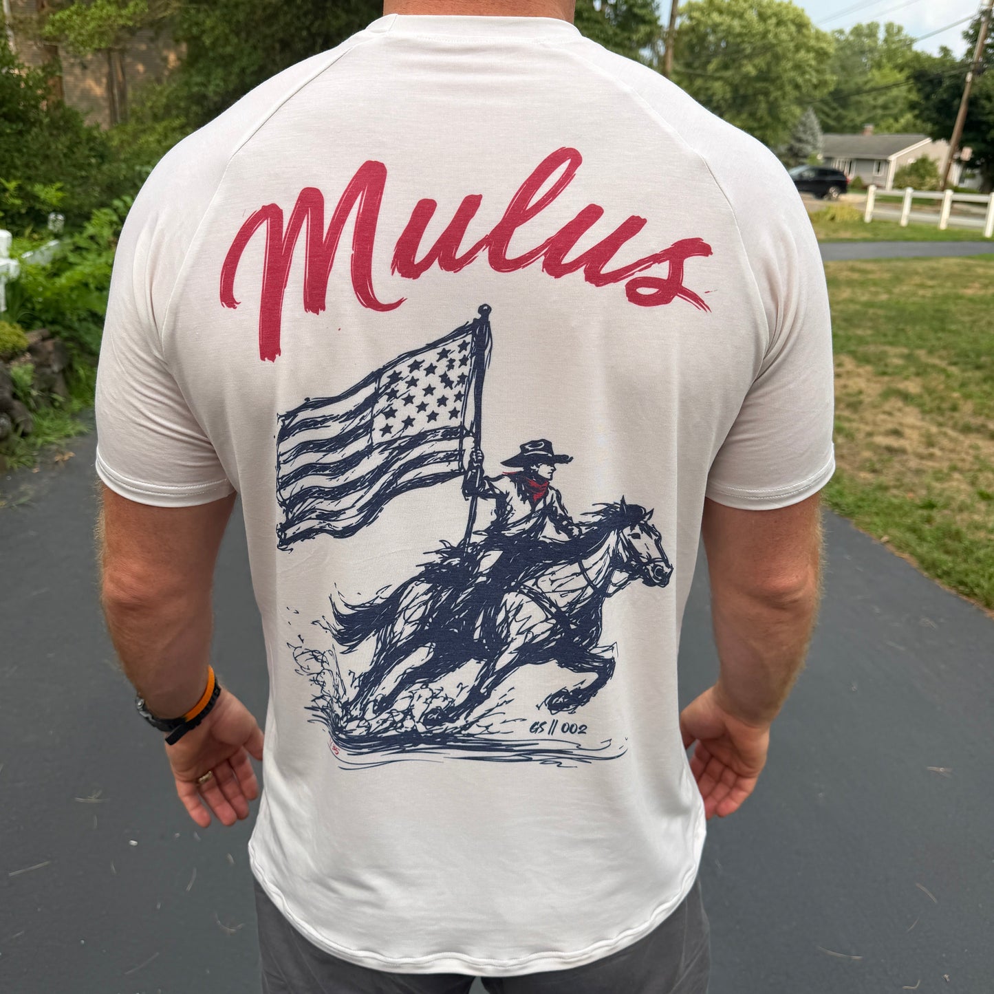 A man viewed from behind wearing a white t-shirt with a large graphic print on the back. The print features the word 'Mulus' in red script and a navy blue illustration of a cowboy riding a horse while holding an American flag.
