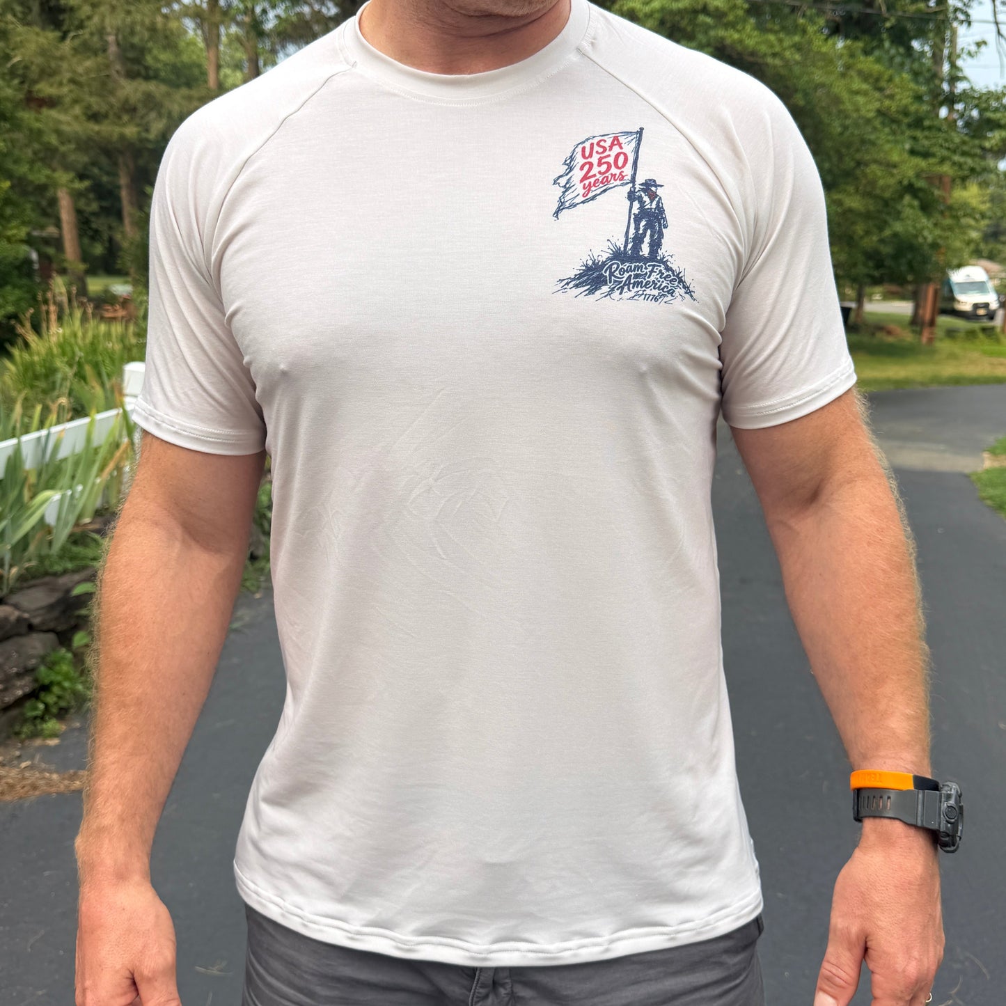 A man wearing a white short-sleeve athletic t-shirt with a blue and red graphic on the left chest, paired with gray athletic shorts, standing outdoors on a paved path.